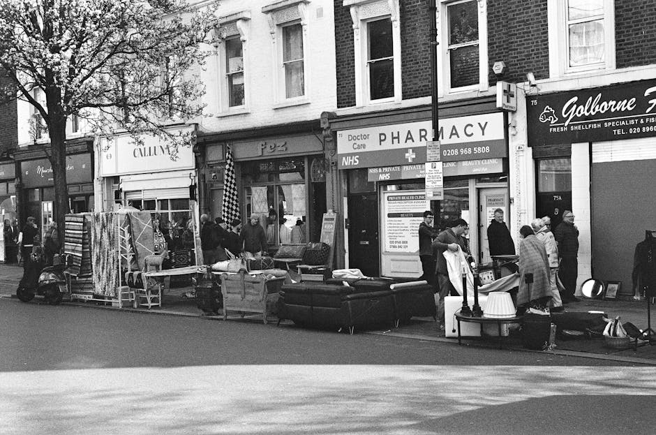 A black-and-white street scene on Barnes High Street SW13 featuring a variety of parked and moving vehicles, including a scooter, with multiple pedestrians walking along the sidewalk. The storefronts visible include a pharmacy with signage advertising NHS and private healthcare services, a fish and seafood shop branded as 'Goringe', and other small retail establishments. The scene shows furniture and other items displayed outside some shops, and a street lamp is positioned near the center of the image. The area appears lively and well-maintained, with trees partially framing the storefronts and typical urban street lighting. The overall setting suggests a busy, commercial environment consistent with central Barnes, with a focus on community activity and local businesses. Carpet Cleaners Barnes emphasizes thorough surface cleaning and sanitisation, aligning with the area's white-glove approach to maintaining hygiene and cleanliness in retail and public spaces.