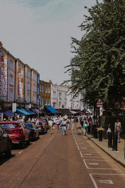 A busy street scene on Barnes High Street SW13, showing a row of colorful, multi-story shops with awnings and signs, lined along a narrow asphalt road. Parked cars are visible along the curb, while pedestrians walk along the sidewalk and cross the street. The area is shaded by large trees, and the buildings feature a mix of brick and painted facades. The scene is well-lit with natural daylight, suggesting a clear day. The image emphasizes a lively commercial area that may benefit from professional cleaning services provided by Carpet Cleaners Barnes for maintaining cleanliness, hygiene, and presentation of the commercial environment.
