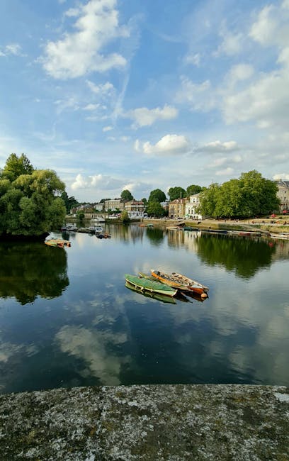 A serene view of residential houses along the riverbank in Barnes, featuring multi-story brick and Victorian-style homes with large windows, surrounded by lush green trees and shrubbery. The clear blu